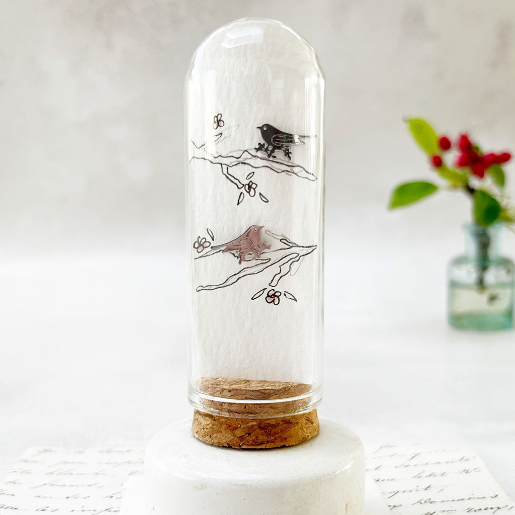 Zamsoe bird jewellery displayed with a hand-drawn illustration in a glass dome
